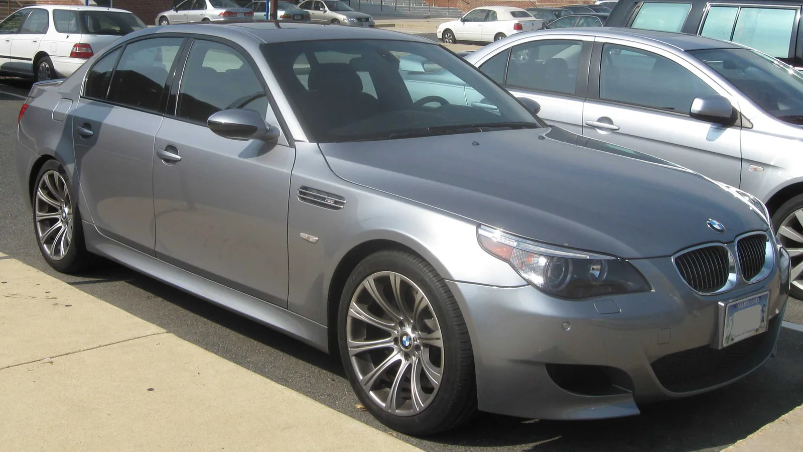 Silver BMW M5 E60 sedan parked outdoors among other cars, front three-quarter view.